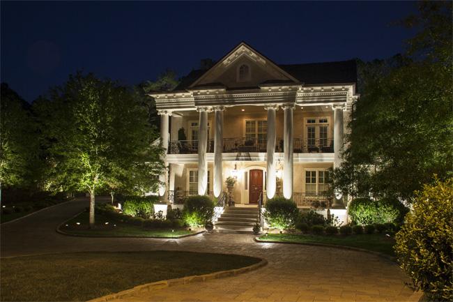 White Plantation-style home with two floors lit up at night with porch lighting and in front yard landscaped plants.