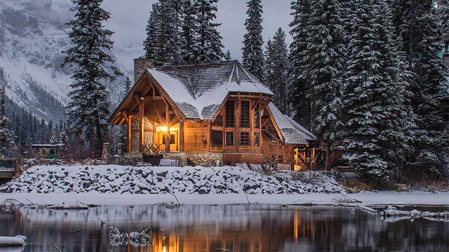 A snowy wooden house in dim midday light covered in snow in front of a winter lake.