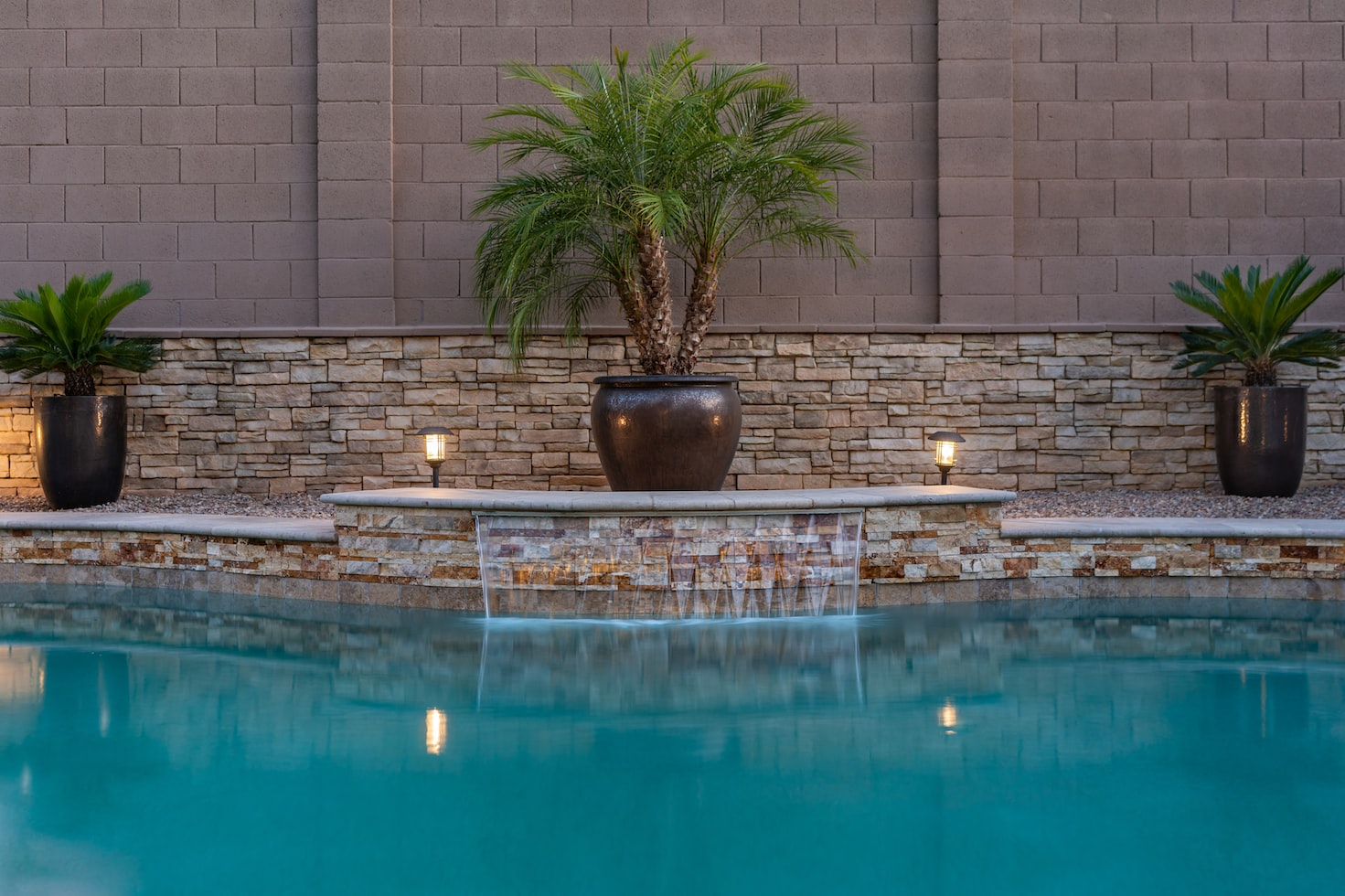 three-palm-trees-in-brown-planters-poolside-nex-to-stone-wall