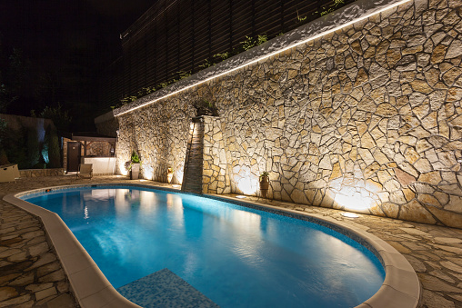 Dimly-lit light blue pool at night with tall rock wall illuminated.