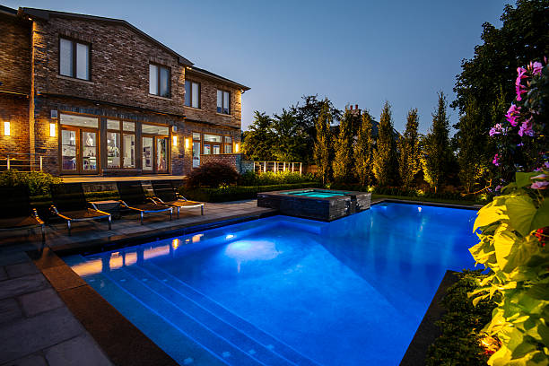 Blue-lit pool at dusk next to stone house with trees and plants and purple flowers in background.
