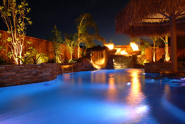 Deep blue- lit pool with patio greenery and plants and hut illuminated.