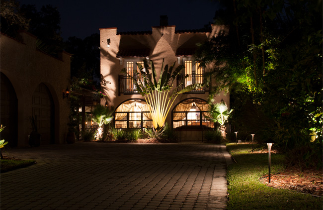 Spanish-style home with tall palms illuminated in front of the house with smaller lighting along stone driveway.