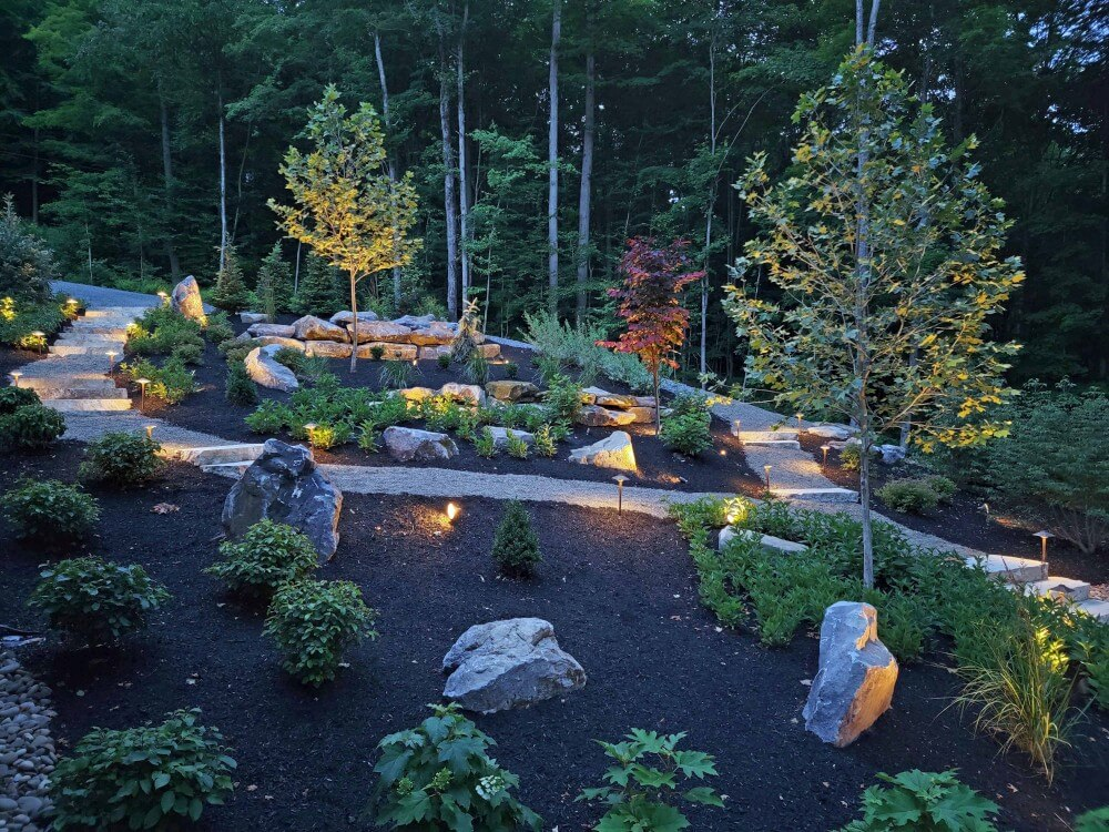 intricate landscaping with bushes, shrubbery, trees, and stone pathway that are all illuminated by path lights